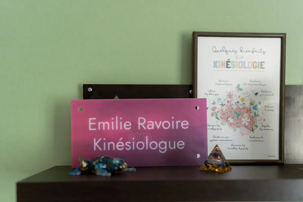 Plaque rose « Émilie Ravoire Kinésiologie » posée sur une étagère à côté de son diplôme encadré
